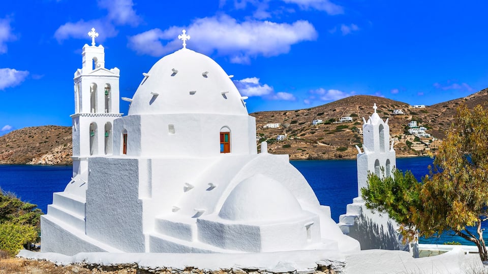 A white church with two towers and a dome, surrounded by a coastal landscape and mountains.