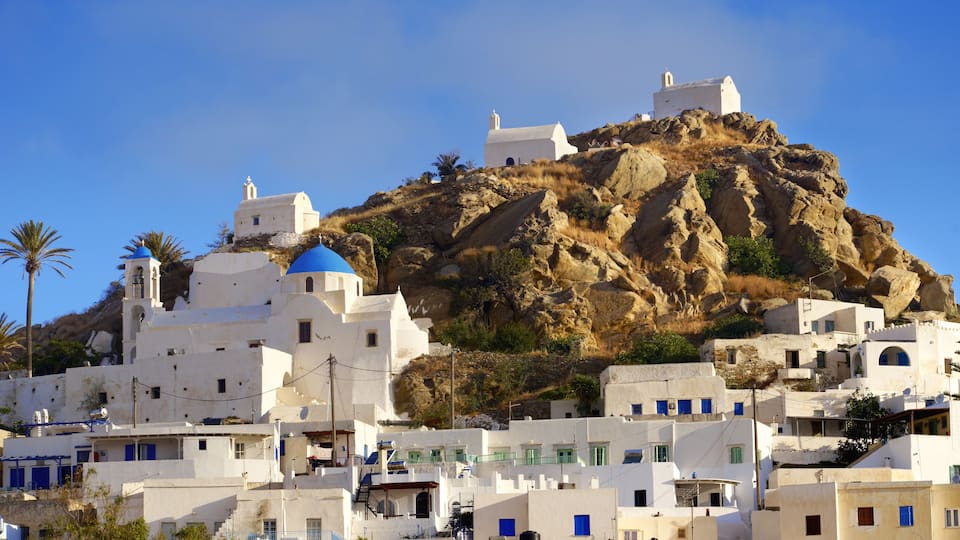 A coastal village with white buildings and blue domes, set against a rocky hillside under a clear blue sky.