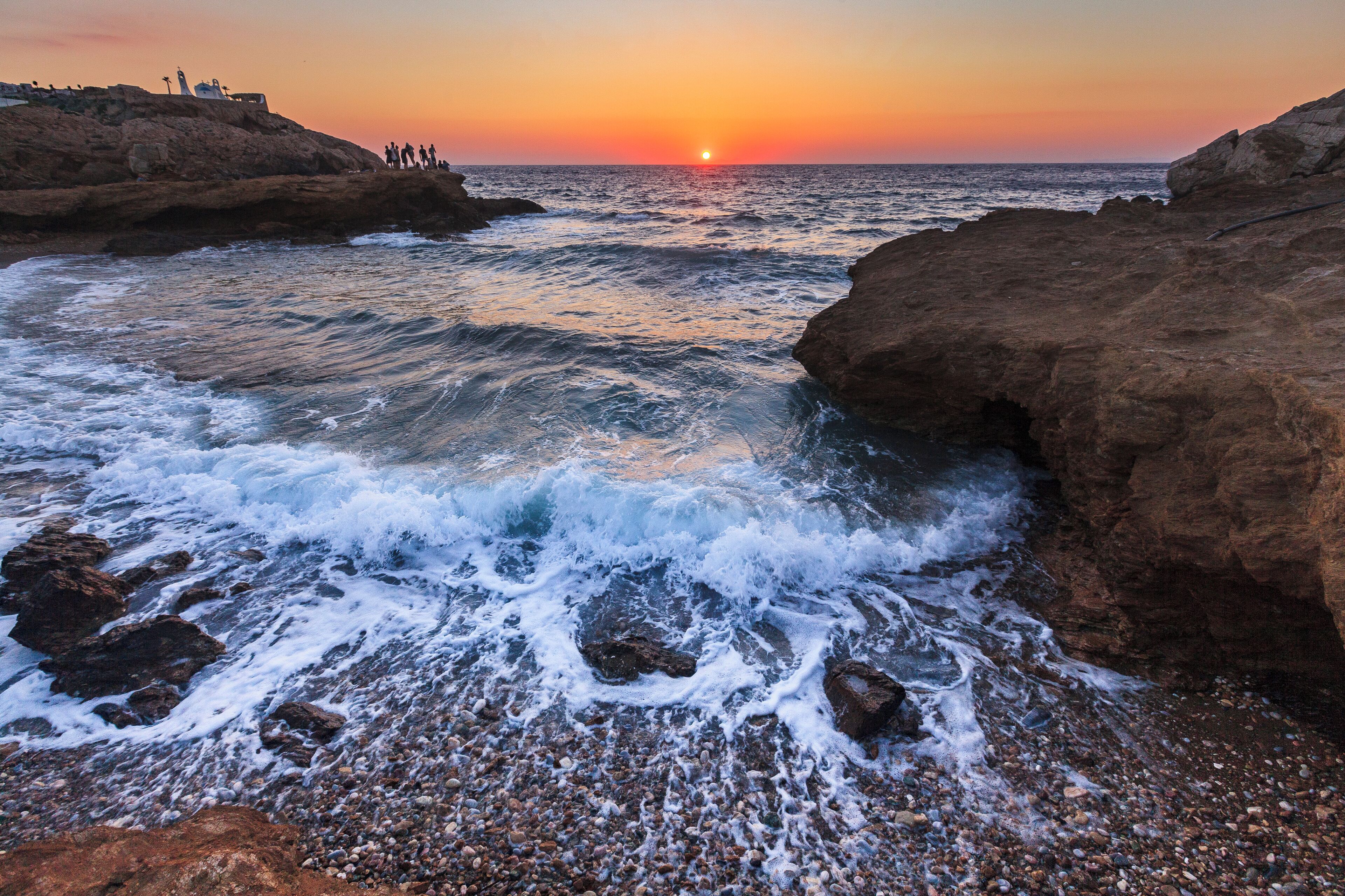 A rocky shoreline with waves crashing against the rocks during sunset.