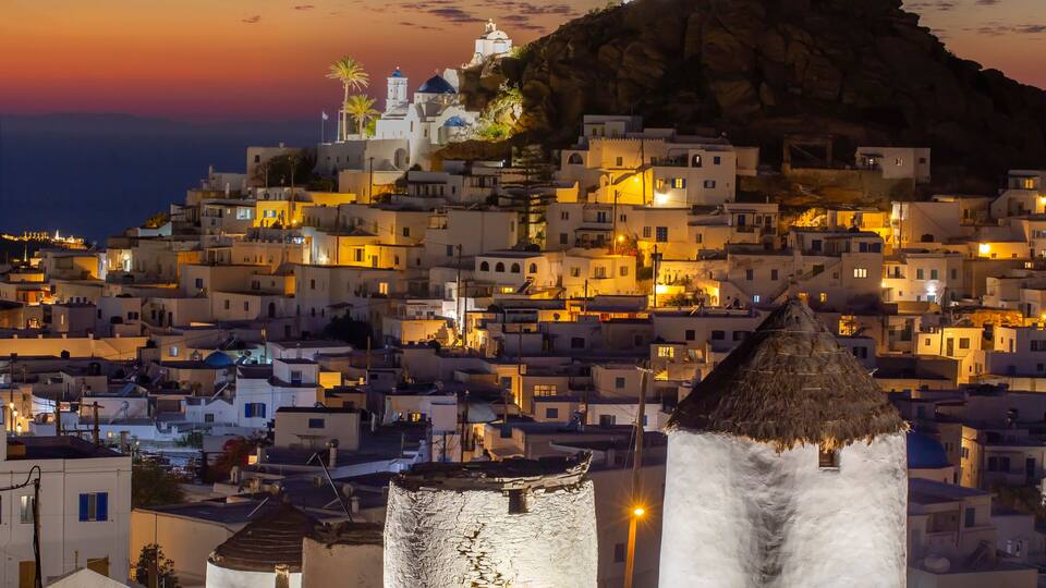 A coastal town at sunset with white buildings, a prominent hilltop church, and a lighthouse.