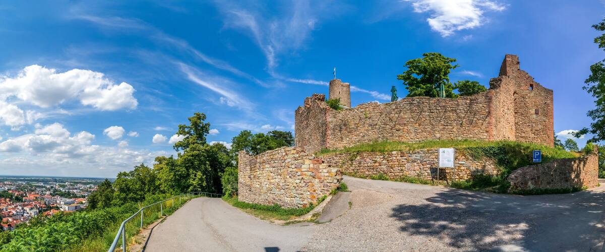 A historic stone castle with a paved road leading up to it, surrounded by lush greenery and a view of a town in the distance.