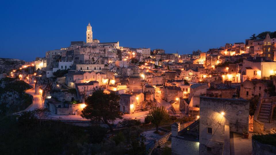 A historic town at dusk with illuminated buildings, a prominent tower, and winding streets.