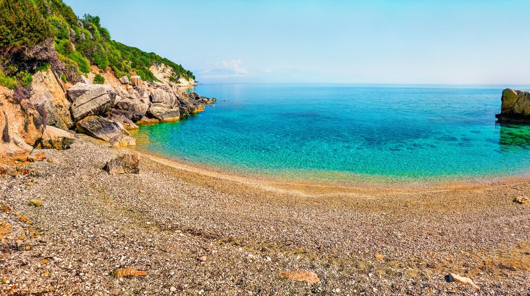 A pebble beach with clear turquoise water, rocky cliffs, and a calm sea.
