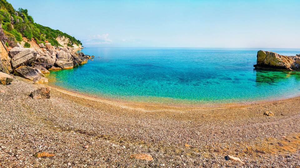 A pebble beach with clear turquoise water, rocky cliffs, and a calm sea.