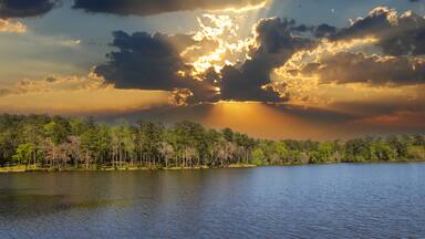 Un lago con una orilla boscosa y un cielo dramático con rayos de sol atravesando las nubes.