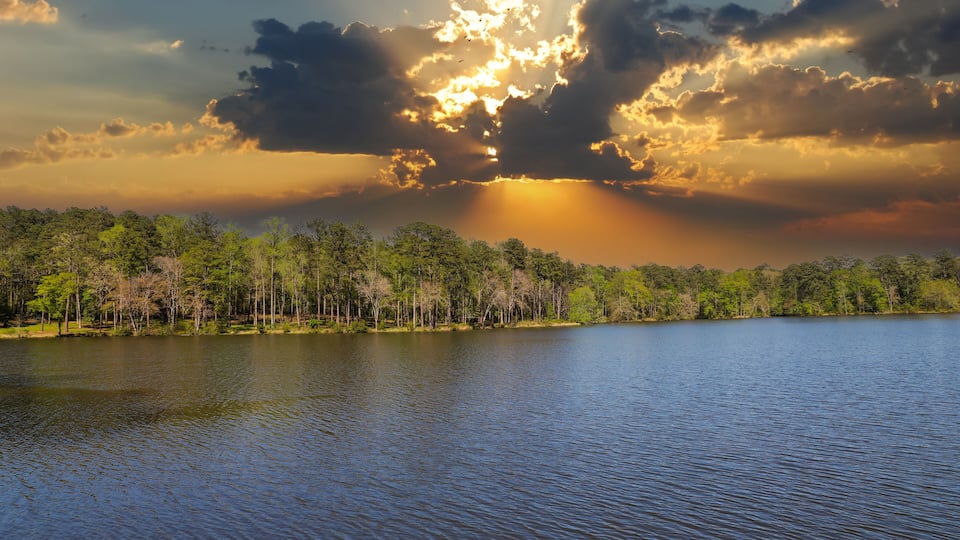A lake with a forested shoreline and a dramatic sky with sun rays piercing through clouds.