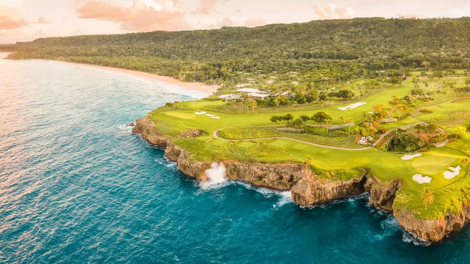A coastal golf course with a view of the ocean and a mountainous landscape in the background.