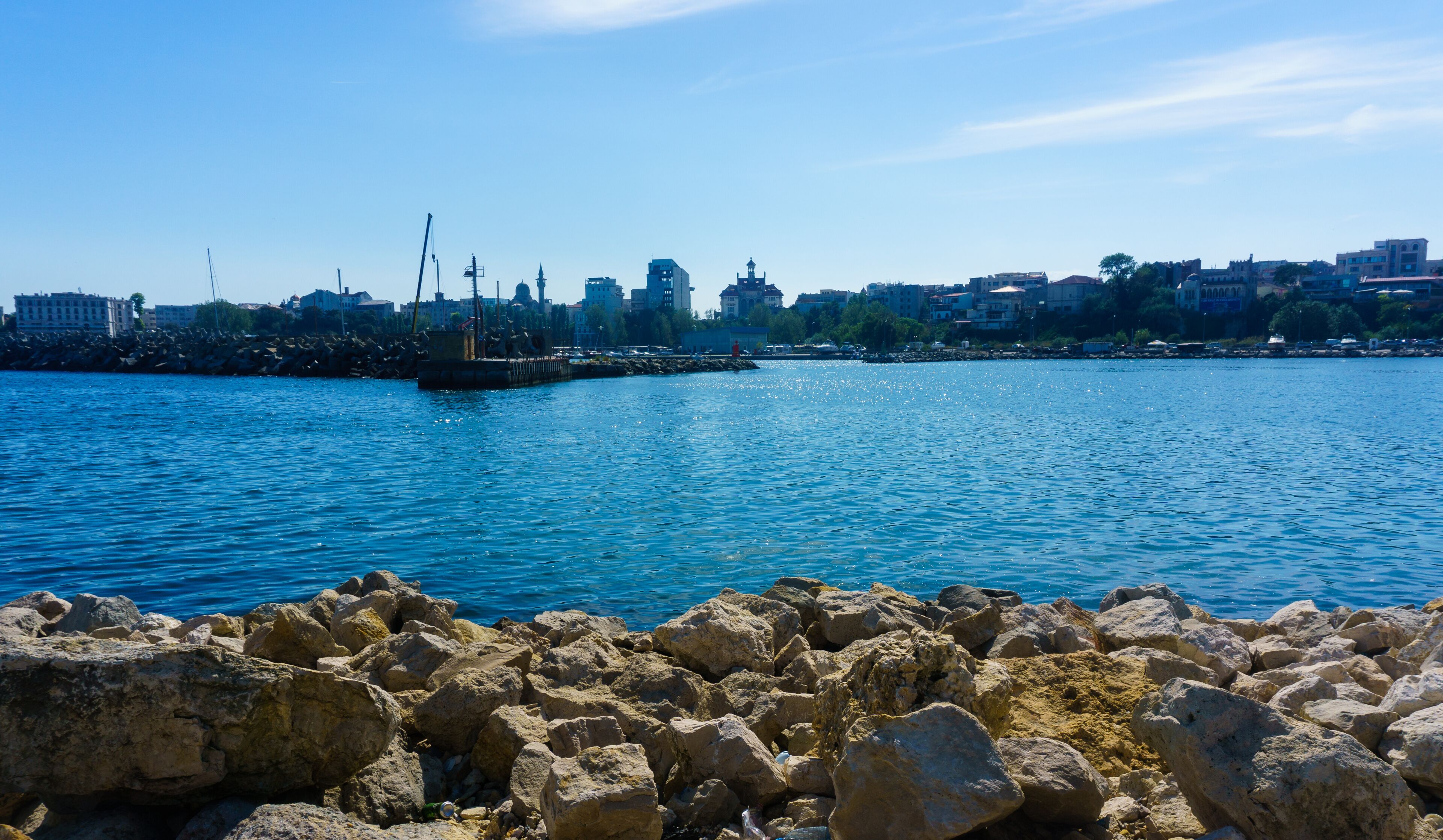 A rocky shoreline with a clear blue sea, a distant city skyline, and a pier with a ship.