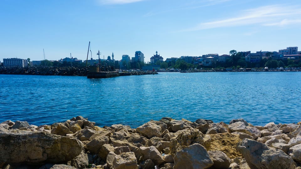 A rocky shoreline with a clear blue sea, a distant city skyline, and a pier with a ship.