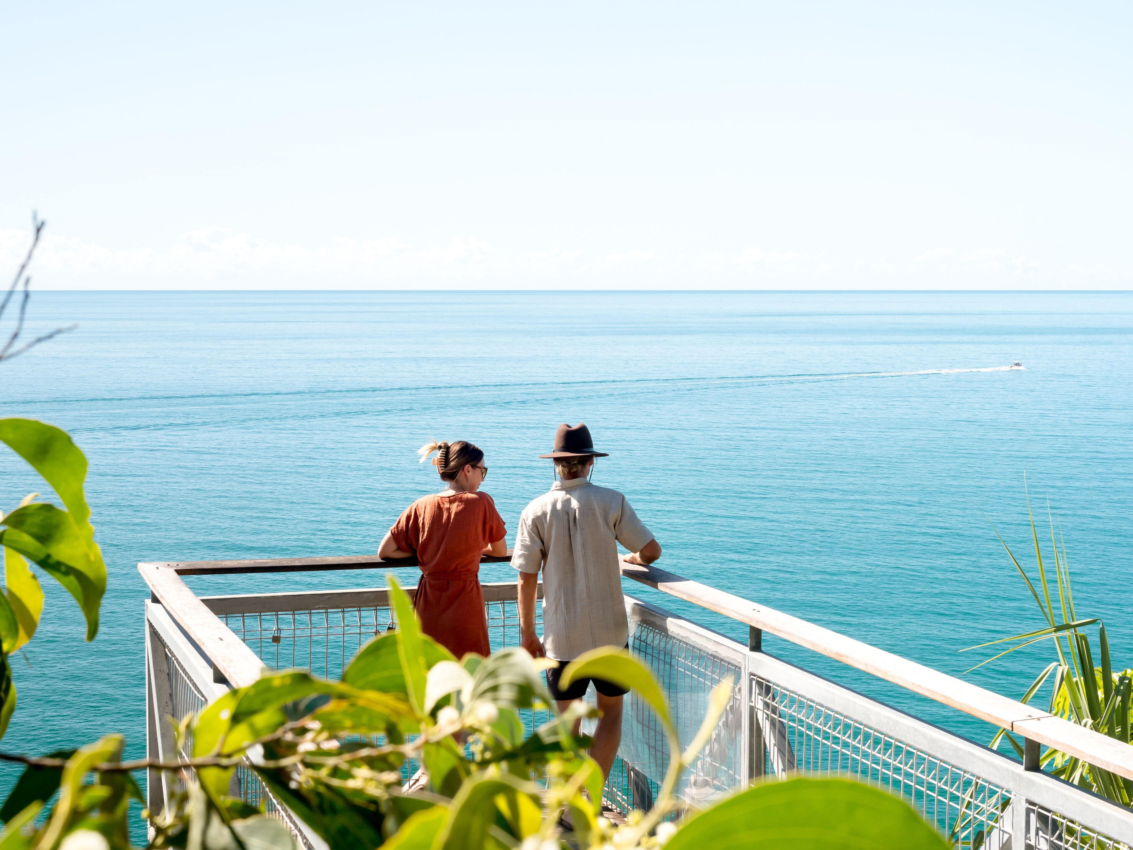 A couple overlooking a calm sea from a balcony with a railing and greenery in the foreground.