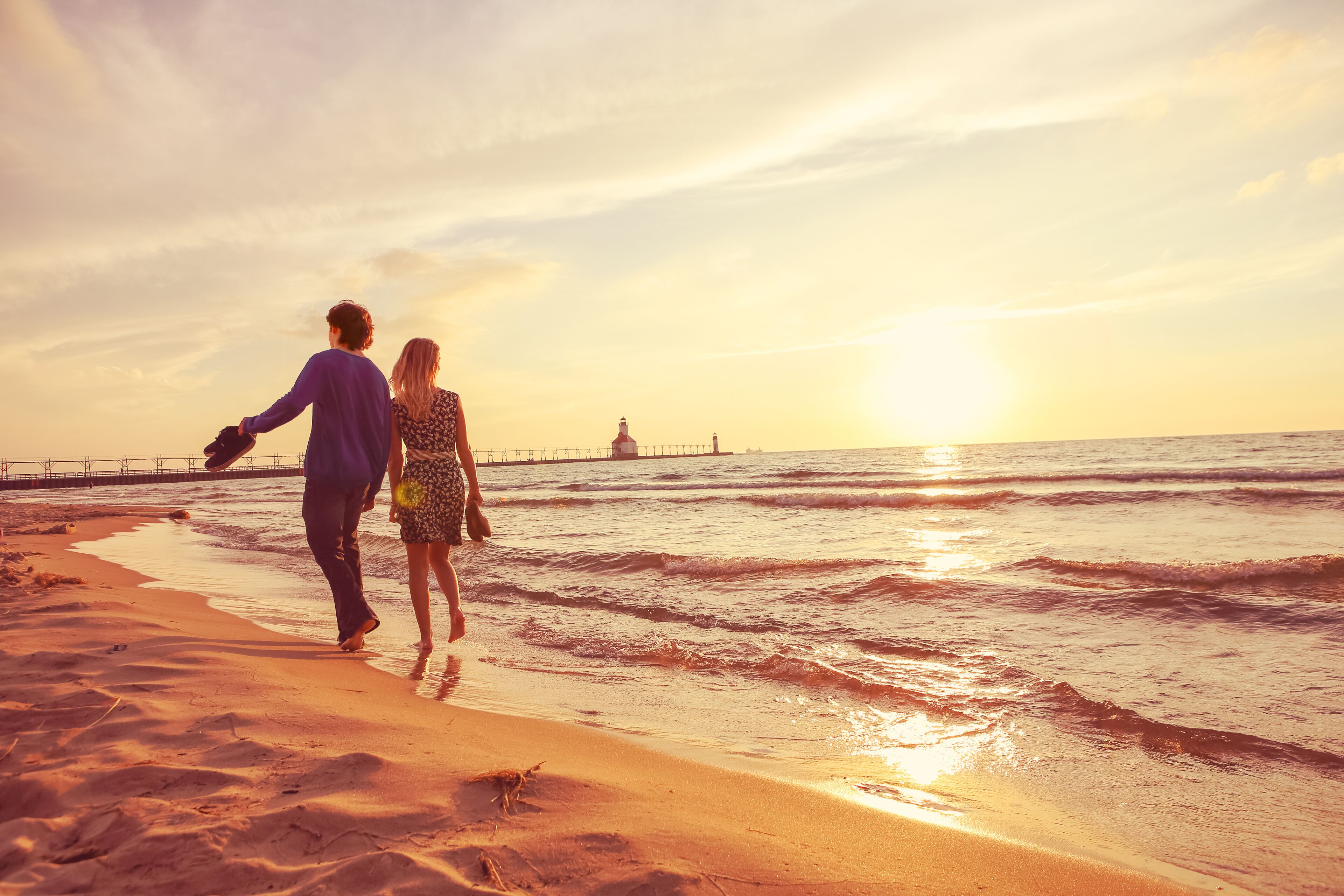 A couple walking on a beach with the ocean and a pier in the background during sunset.