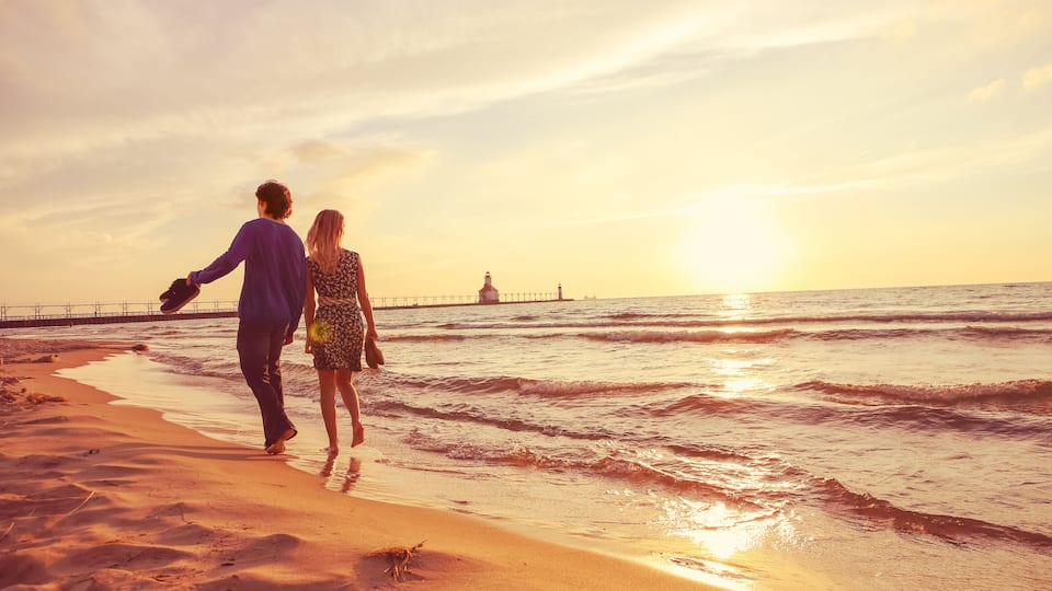 A couple walking on a beach with the ocean and a pier in the background during sunset.