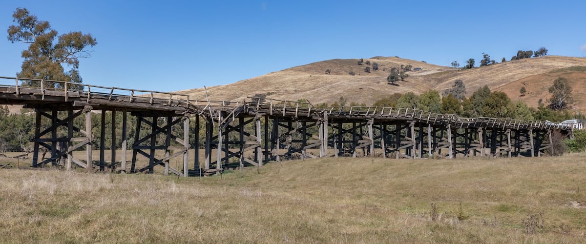 A wooden bridge over a dry grass field with a hilly landscape in the background.