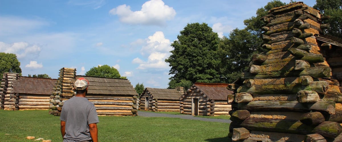 A man standing in front of a log cabin with a clear sky and trees in the background.