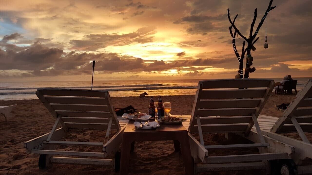A beach with two lounge chairs, a table with food and drinks, and a sunset in the background.