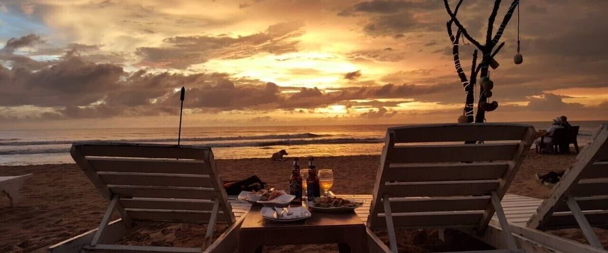 A beach with two lounge chairs, a table with food and drinks, and a sunset in the background.