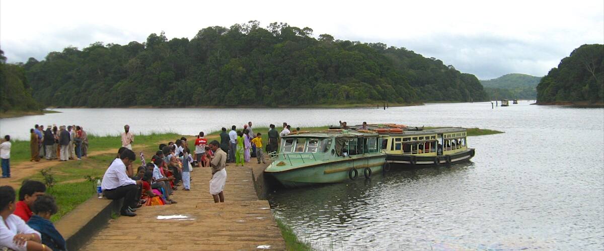 A group of people on a dock by a river, with boats in the water and a forested hillside in the background.
