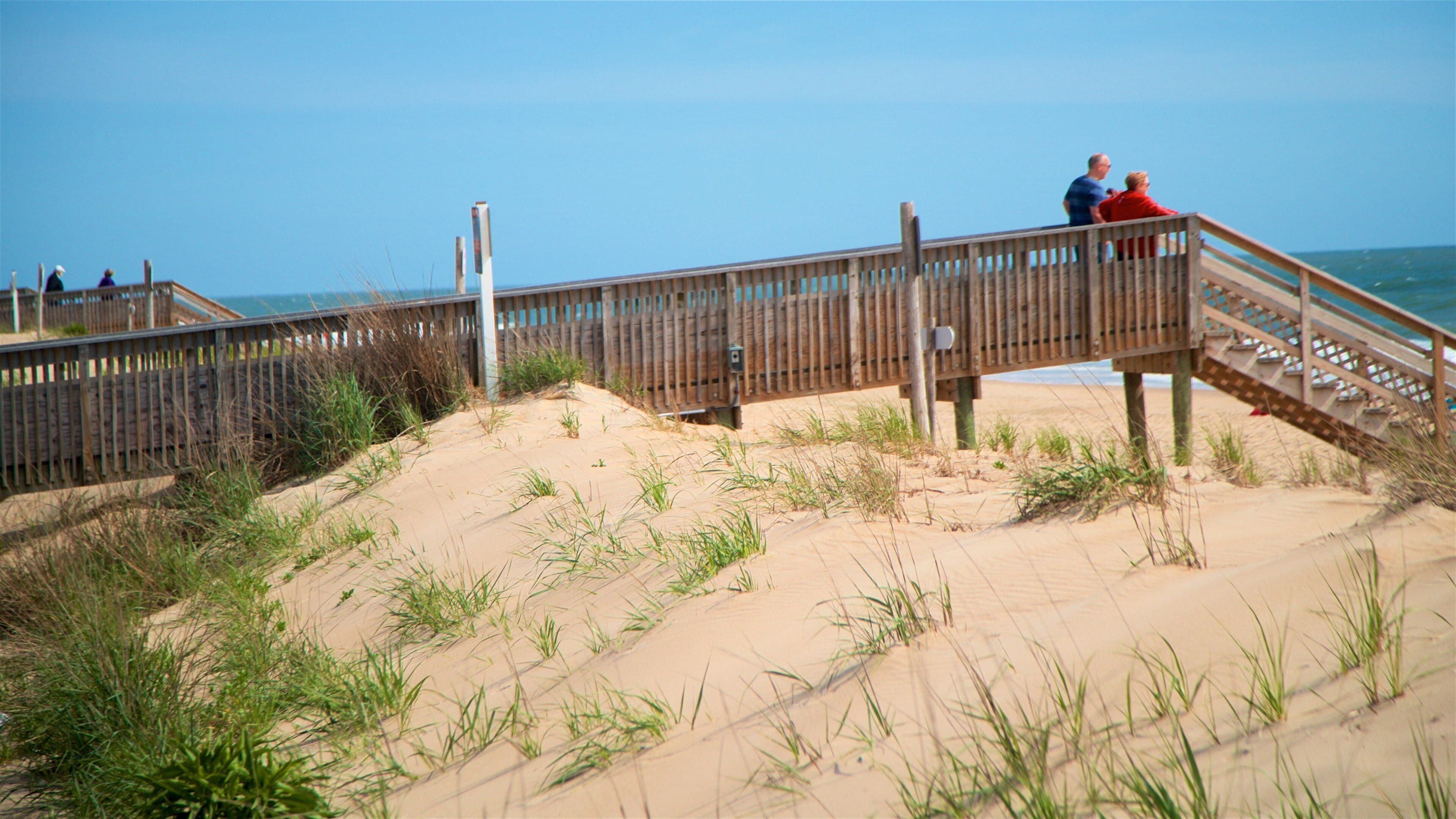 A wooden boardwalk leading to a beach with sand dunes and grass.