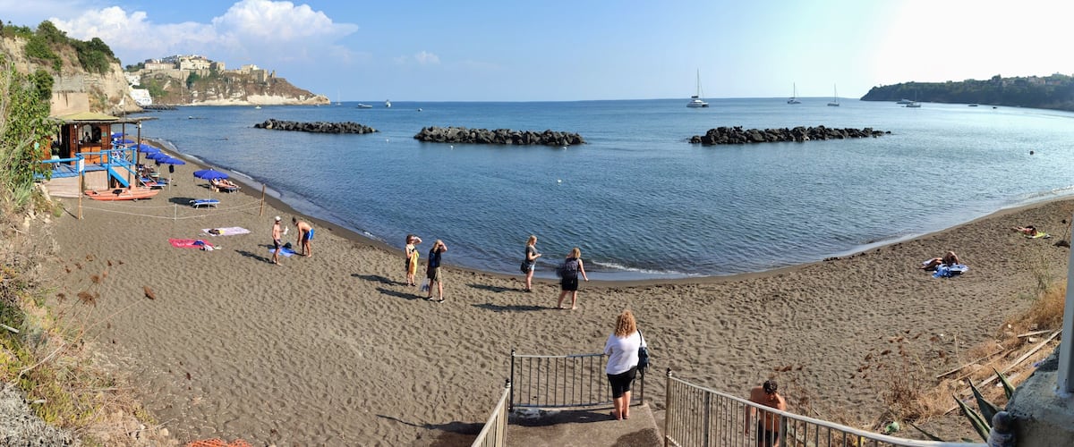 A beach with people sunbathing, a rocky outcrop, and boats in the water.