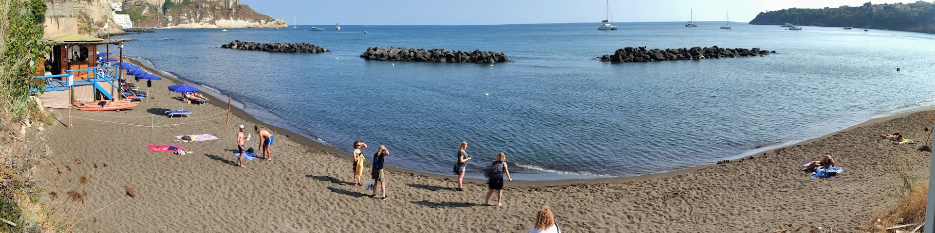 A beach with people sunbathing, a rocky outcrop, and boats in the water.