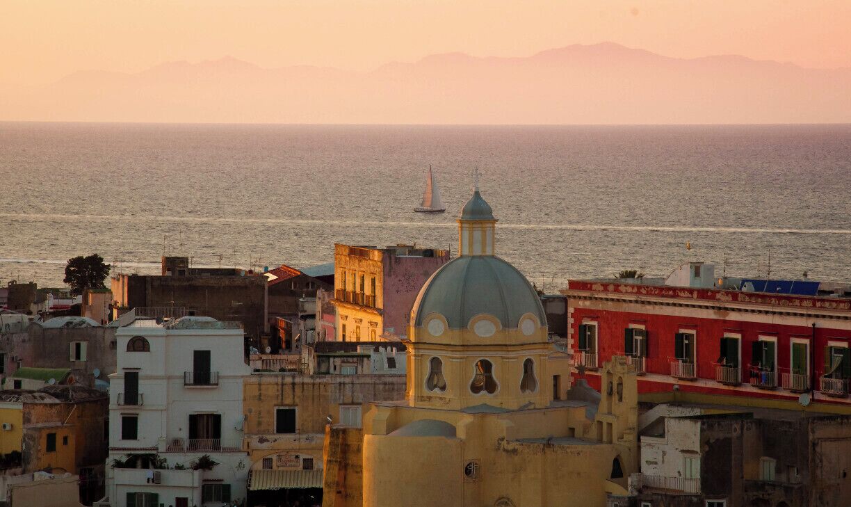 A coastal cityscape with a prominent church dome, colorful buildings, and a sailboat on the sea.