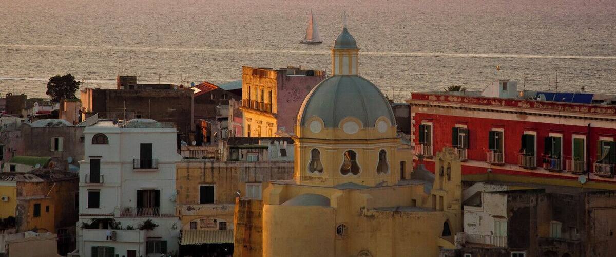 A coastal cityscape with a prominent church dome, colorful buildings, and a sailboat on the sea.