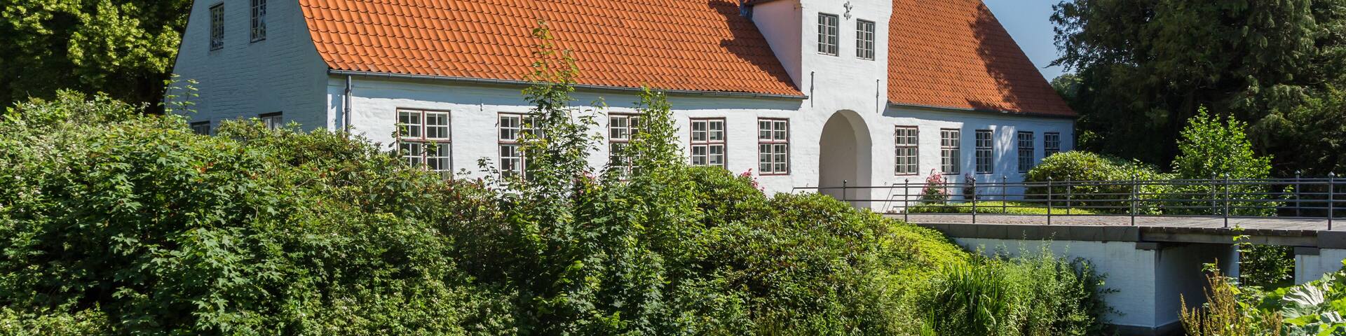 A white building with a red-tiled roof and a small tower, surrounded by greenery and a bridge.