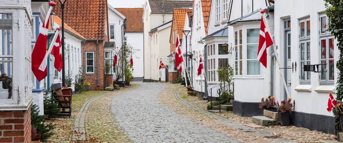 A cobblestone street lined with white buildings, red flags, and potted plants.