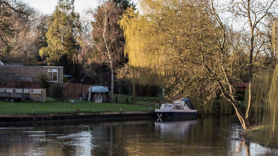 A calm river with a boat docked on the left, surrounded by trees and a grassy area with a small building.