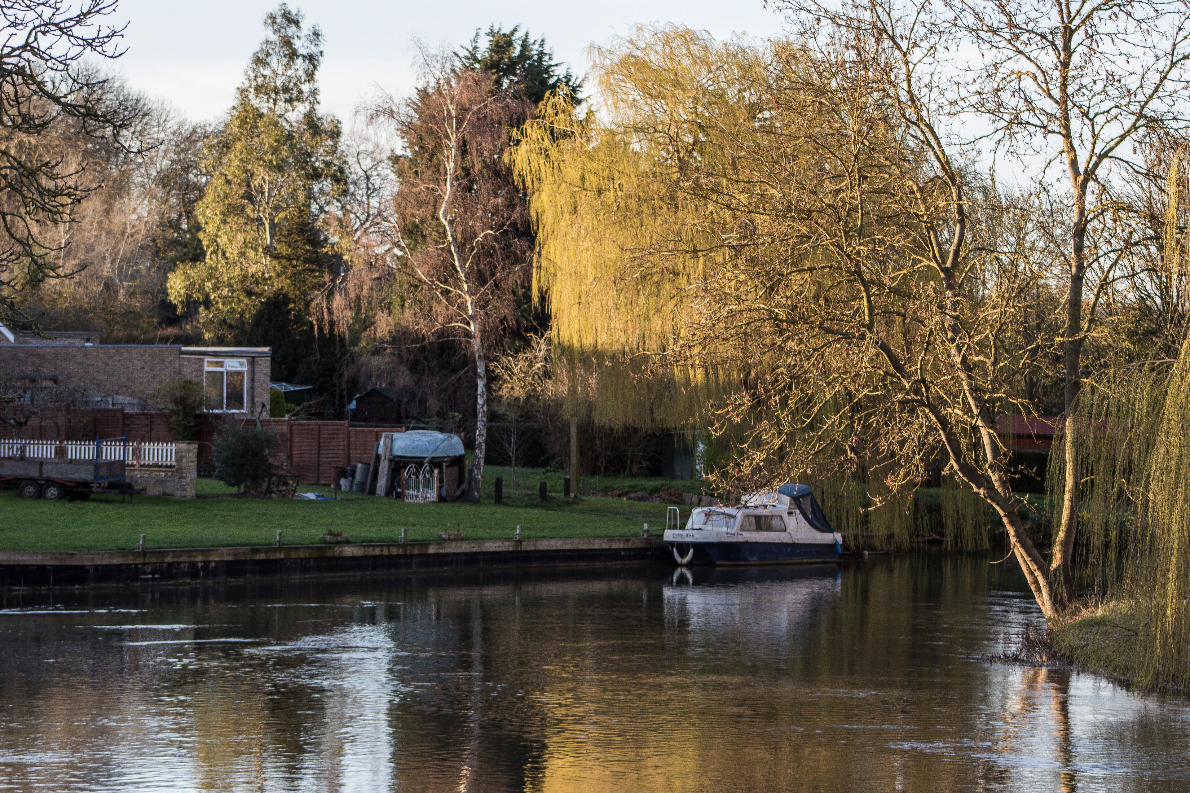 A calm river with a boat docked on the left, surrounded by trees and a grassy area with a small building.