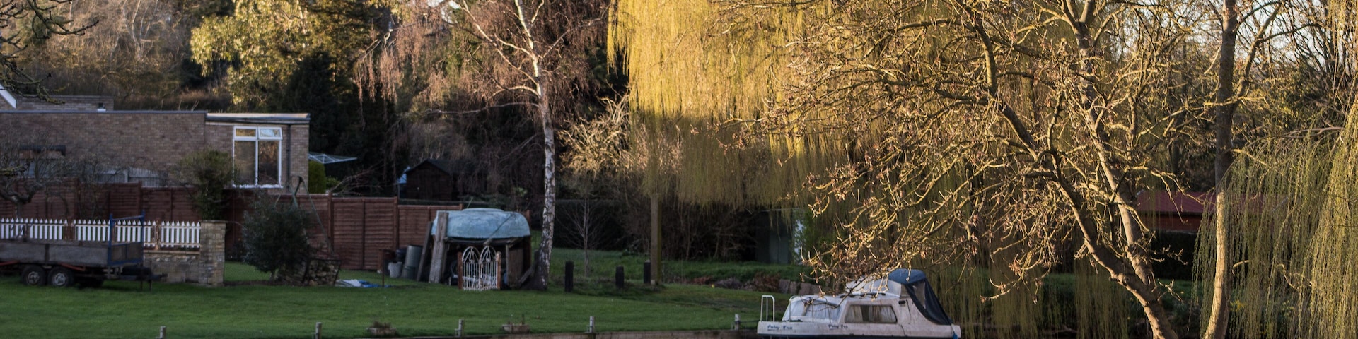 A calm river with a boat docked on the left, surrounded by trees and a grassy area with a small building.
