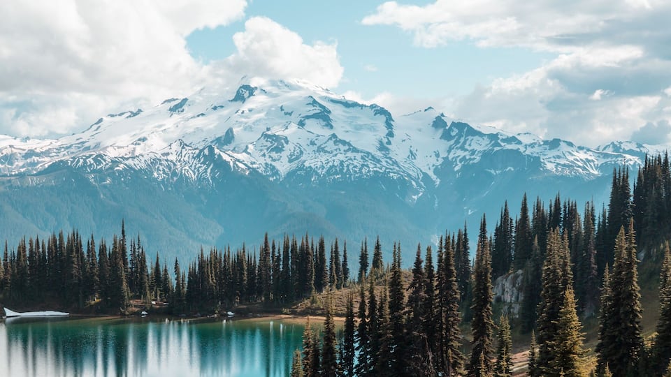 A mountain range with snow-capped peaks, a turquoise lake, and dense coniferous forests.