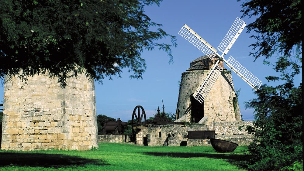 A historic windmill with a large sail, surrounded by stone walls and greenery.
