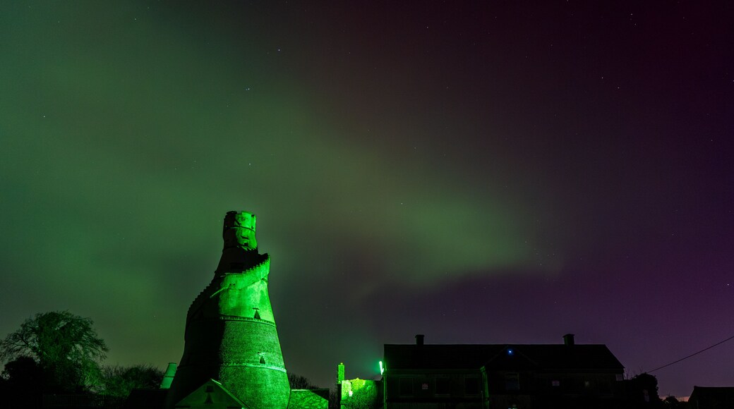 A green-lit tower with a conical roof, surrounded by buildings and a grassy area under a night sky with visible stars.