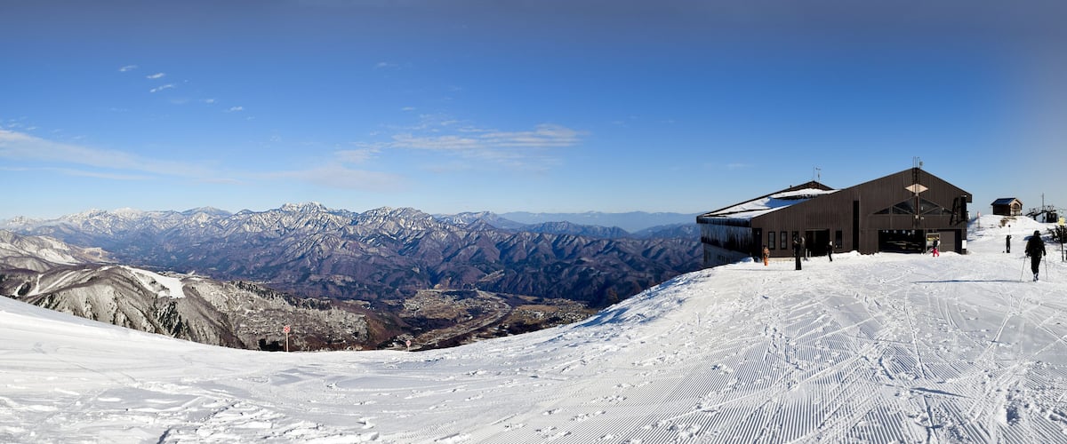 A snowy mountain slope with ski tracks, a wooden building, and a clear blue sky.