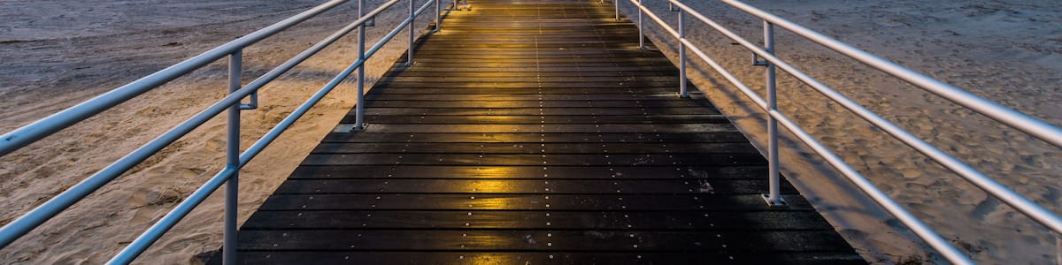 A wooden boardwalk leading to a beach with a railing on both sides and a street lamp at the end.