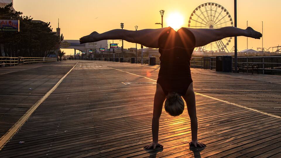 A person performing a handstand on a wooden boardwalk with a sunset and a Ferris wheel in the background.