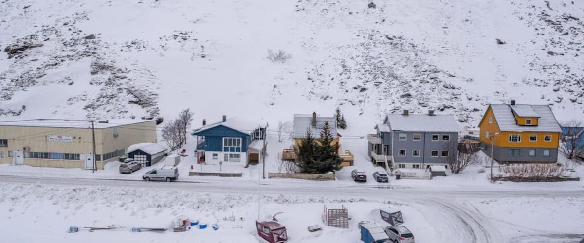 A snow-covered landscape with a mix of residential buildings, a road, and a hill in the background.