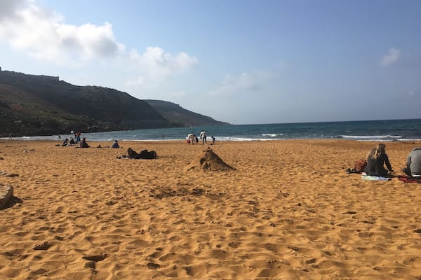 A sandy beach with footprints, a few people, and a hilly landscape in the background.