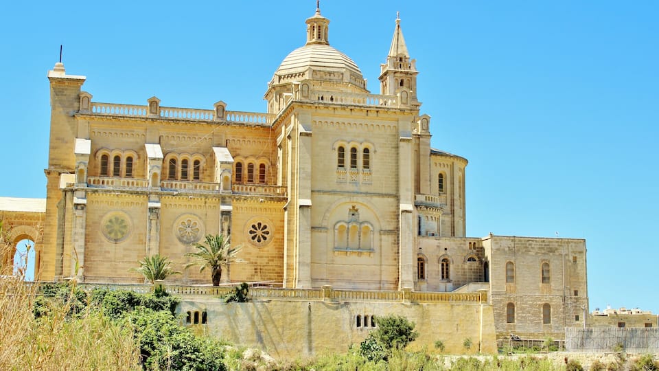 A large, historic cathedral with a prominent dome and bell tower, surrounded by lush greenery.