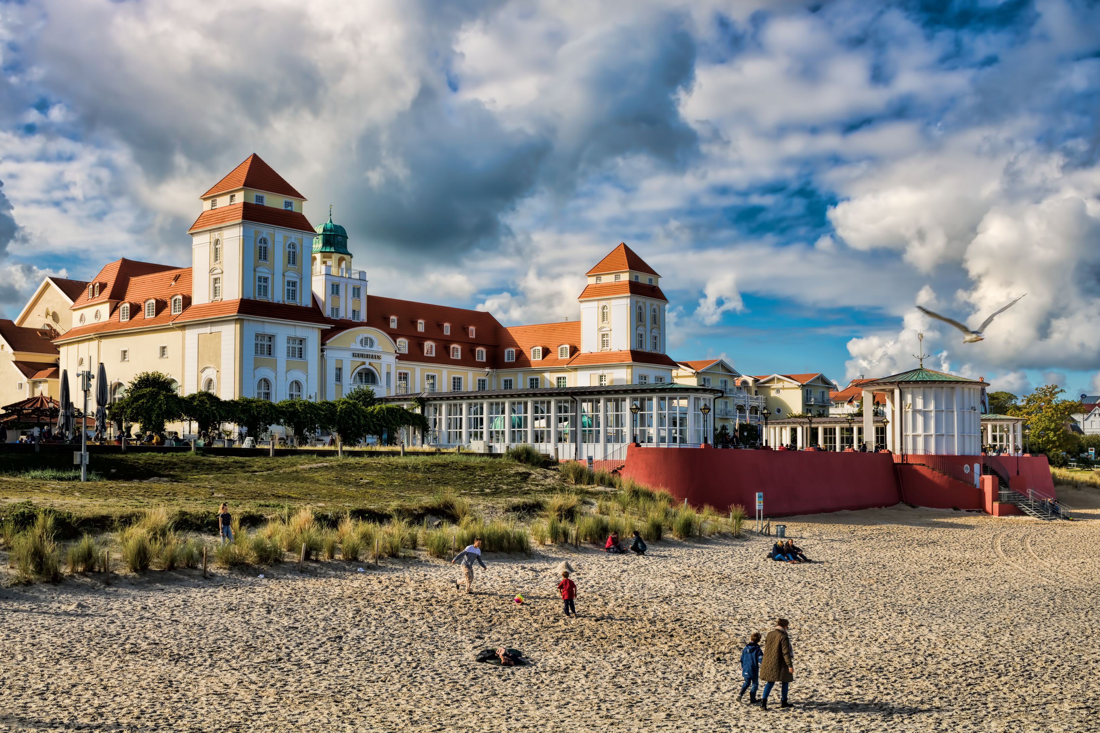 A coastal building with red roofs and a green dome, surrounded by a sandy beach with people and grass.