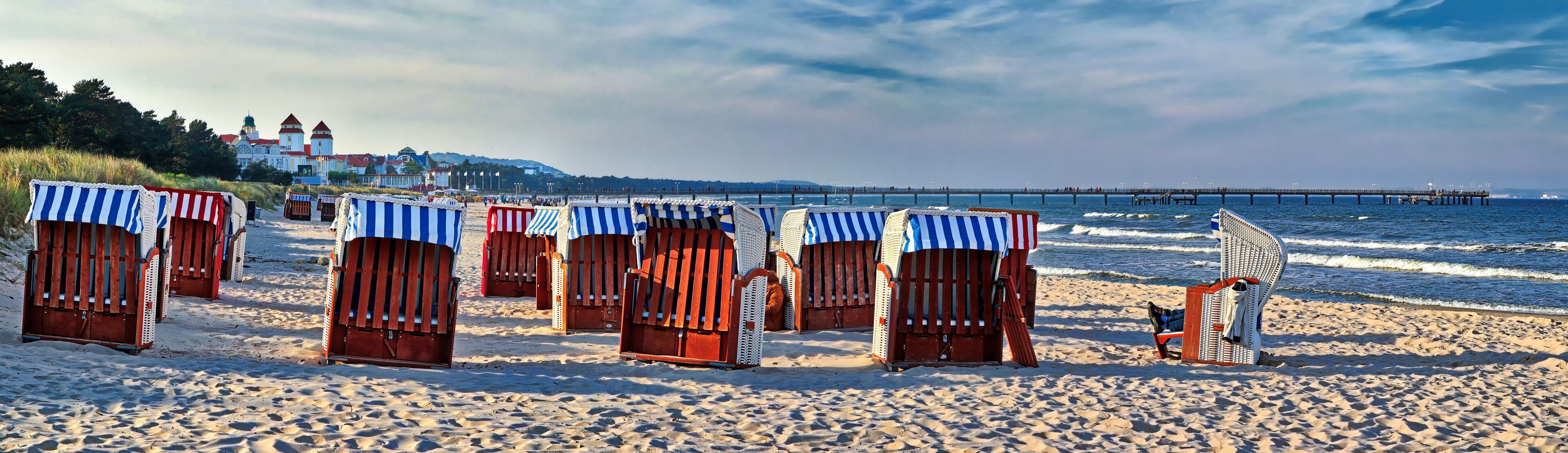 A beach with striped beach huts and a pier in the background.