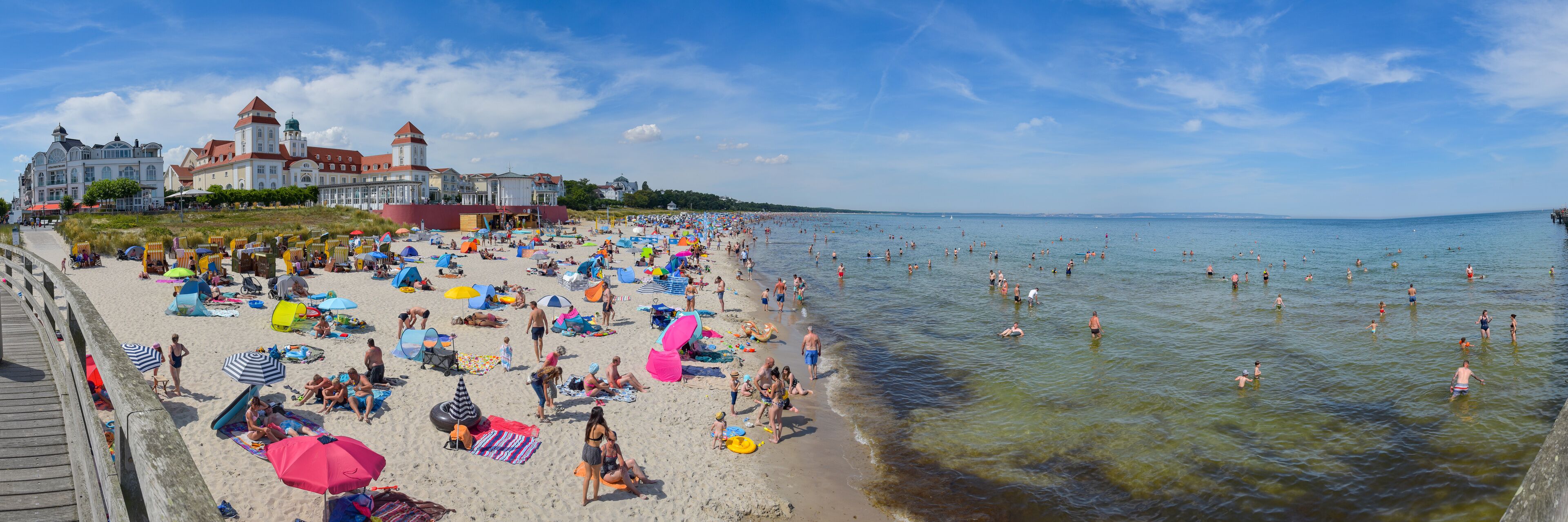 A crowded beach with people sunbathing and swimming, colorful umbrellas, and a historic building in the background.