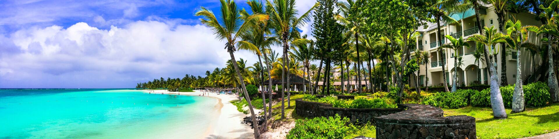 A beach with turquoise water, palm trees, and a resort with a stone wall and steps.
