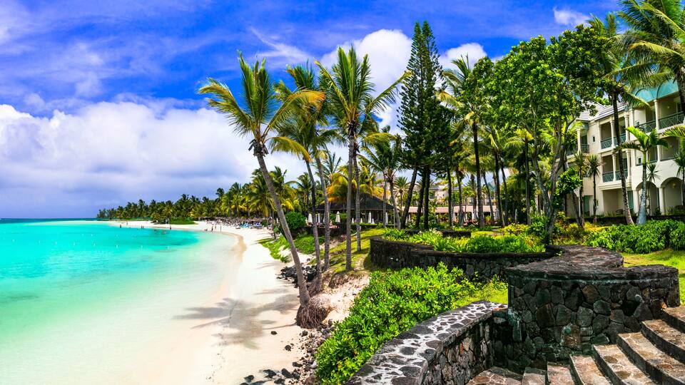 A beach with turquoise water, palm trees, and a resort with a stone wall and steps.
