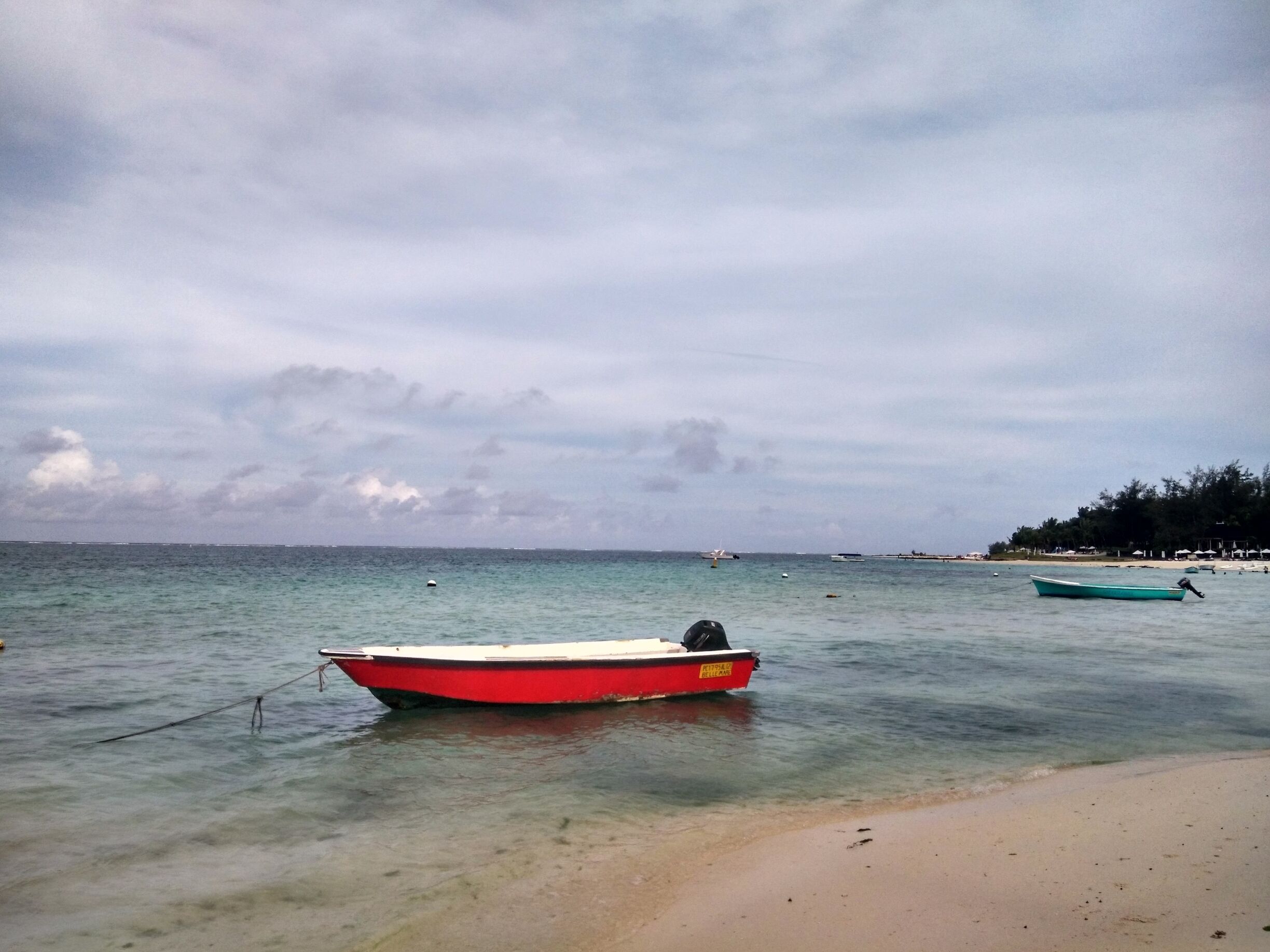 A red boat tied to a buoy on a sandy beach with calm waters and a cloudy sky.