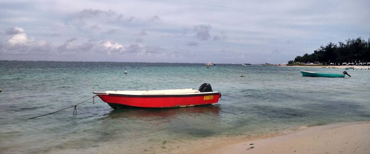 A red boat tied to a buoy on a sandy beach with calm waters and a cloudy sky.