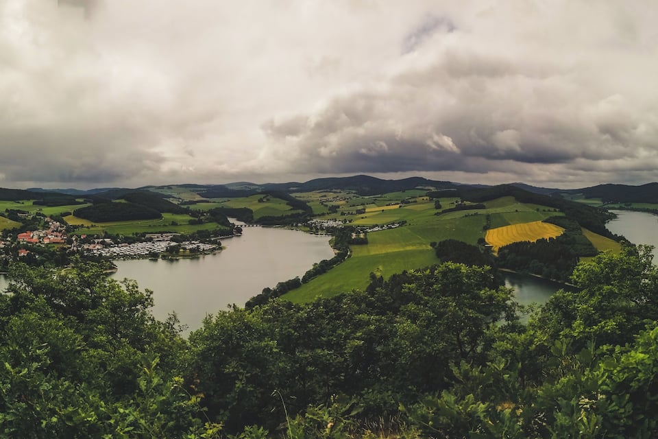 A river winding through rolling hills, a small town with red-roofed buildings, and a cloudy sky.