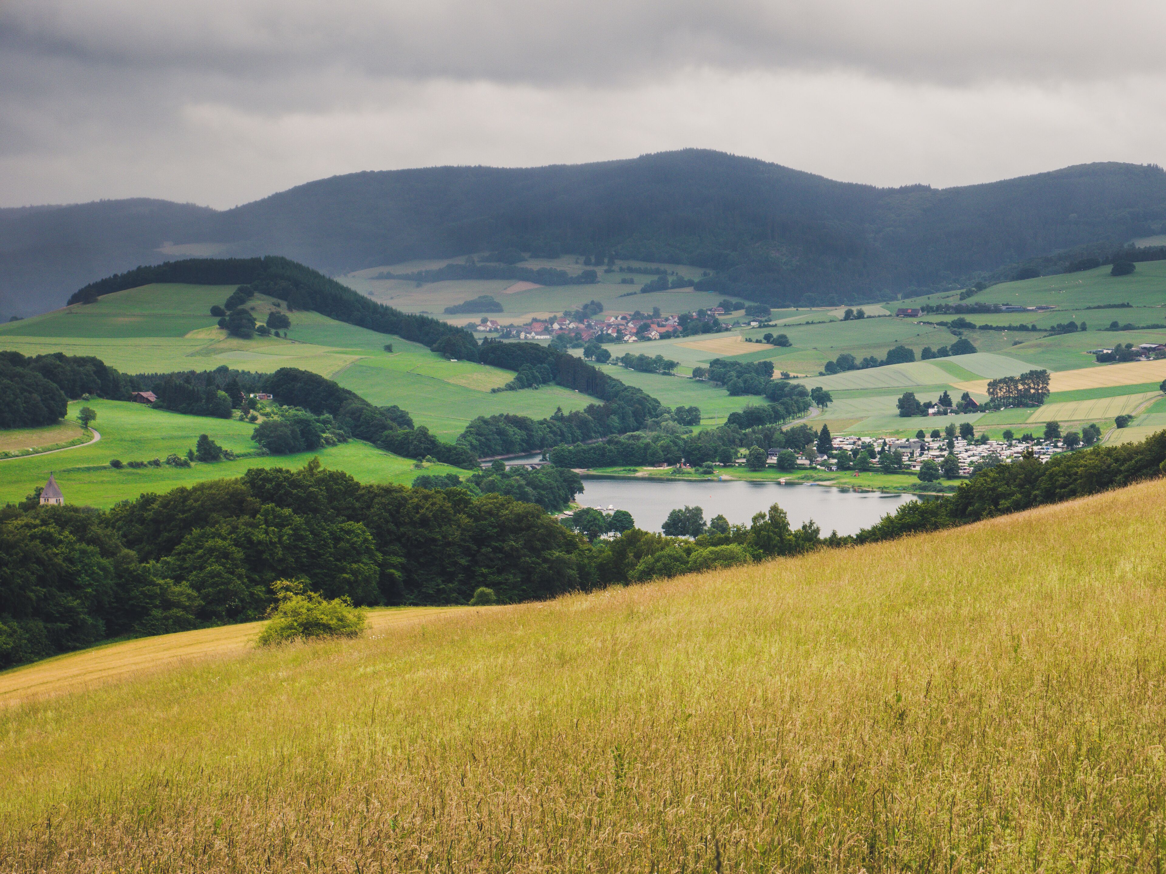 Rolling green hills, a small village, and a lake surrounded by mountains.
