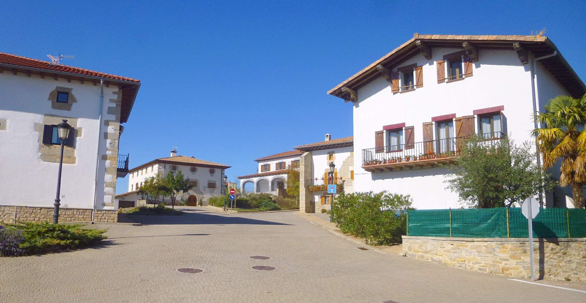 A sunny street in a European village with white buildings, a clear blue sky, and a green fence.